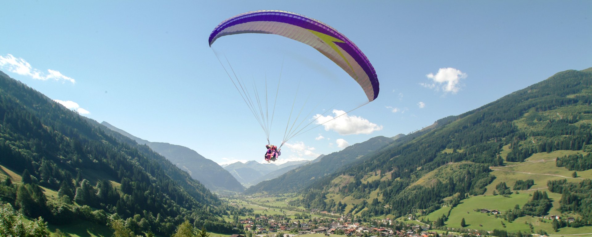 Paragleiter schwebt über Gastein, vor ihm ein Panorama aus grünen Almen, Wäldern und Bergen.